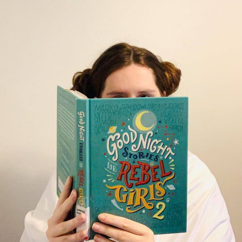 Shayla, a light skinned woman with reddish brown hair, reads Good Night Stories for Rebel Girls 2. Her hair is up in Princess Leia buns and she's wearing a white robe.